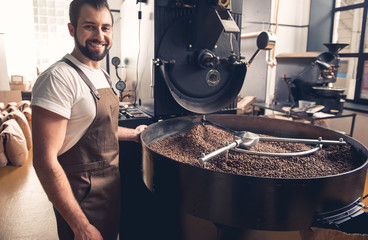 Portrait of happy unshaven male having job at factory. He controlling preparing coffee beans in...