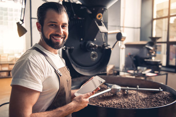 Portrait of cheerful bearded worker using tablet while locating near coffee roaster with spinning cooler professional machine. Industry concept
