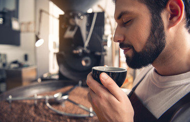 Side view smiling unshaven worker drinking cup of appetizing beverage while locating at factory. Rest concept