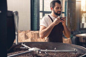 Portrait of cheerful professional bearded coffee roaster drinking delicious hot beverage near appliance during break. Relax concept