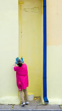 elderly female house painter atiptoe in dark pink overall paints yellow wall with blue downcomer