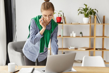 Portrait of female freelancer works remotely at home, uses smart phone for connecting with boss and modern laptop computer, stands at working place near table with papers and cup of hot coffee