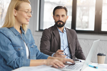 Serious bearded man in jacket and shirt controls female`s work, collaborate together, work on common business presentation on generic laptop computer, sit over office interior. Teamwork concept