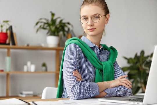 Confident young female employer keeps hands crossed, sits at work place in front of opened laptop, uses free high speed internet connection for searching information online. People, job concept