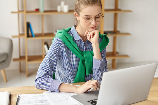 Serious concentrated female student being busy before session, writes course paper in finances, keyboards on laptop, searches necessary information in intrenet, has clever and confident look