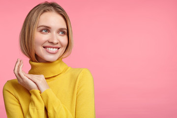 Fototapeta premium Beautiful woman has pleased expression, wears yellow convinient turtleneck sweater, looks dreamfully aside as thinks about something pleasant, isolated over pink studio background with copy space