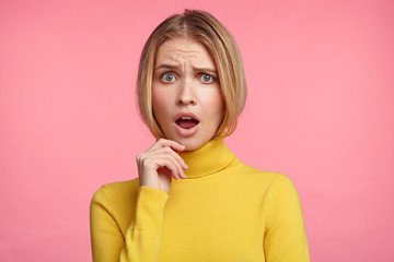 Horizontal portrait of beautiful stupefied woman keeps mouth opened, has upset expression, dressed casually, being confused or worried, sees no way out in difficult situation, poses over pink studio