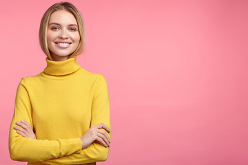 Fototapeta premium Horizontal portrait of smiling happy pleasant looking female wears yellow turtleneck sweater, keeps hands crossed, advertises something, poses on pink background with copy space for promotional text