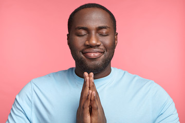 Pleased hopeful strong black man closes eyes, keeps palms together, prays to have good luck, has dreamful look, isolated over pink background. Attractive African American man begs for successful day