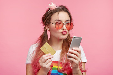 Fashionable female shopaholic chooses new stylish outfit, sees something appealing, rounds lips with great pleasure, holds plastic card, ready to buy it immediately. Woman pays for purchase.