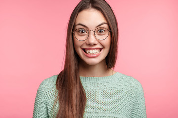 Fototapeta premium Attractive Asian female has funny expression, smiles broadly, hears comic story or anecdote from friend, pretends smiling. Cheerful woman in spectacles has clueless look, isolated on pink wall