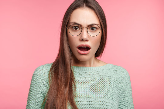 Stupefied shocked woman looks with shocked expression into camera, frowns face in dissatisfaction, astonished to hear shocking news. Female in eyewear has puzzled look, isolated over pink background