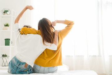 Back view of women lesbian happy couple waking up in morning, sitting on bed, stretching in cozy bedroom, looking through window. Funny women after wakeup. Lesbian couple together indoors concept.