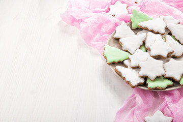 Christmas gingerbread cookies on a plate. Cookies covered with multi colored icing in pale pastel colors shaped as fir-tree and star. White background, pink napkin