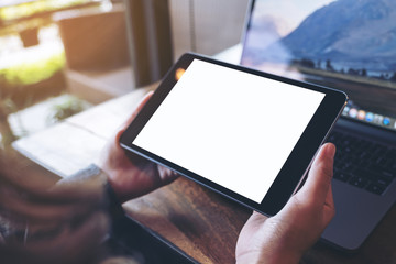 Mockup image of hands holding black tablet pc with white blank screen and laptop on wooden table background in cafe