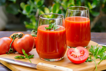Two glasses of fresh tomato juice and tomatoes on a wooden cutting board on green nature background.