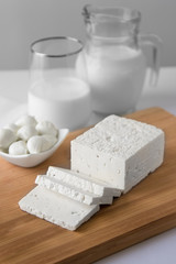 Sliced fresh white cheese and mozzarella balls with glass with milk on wooden board on white background in high key.