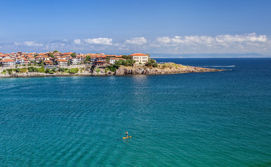 Landscape with the view of the part of old Sozopol town surrounded by the sea. There is an unrecognizable man on the stand-up paddle board for surfing in the sea. Bulgaria, the Black Sea coast.