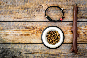Toys for dog stick and ribbon bone near collar and dry food in bowl on wooden background top view copyspace