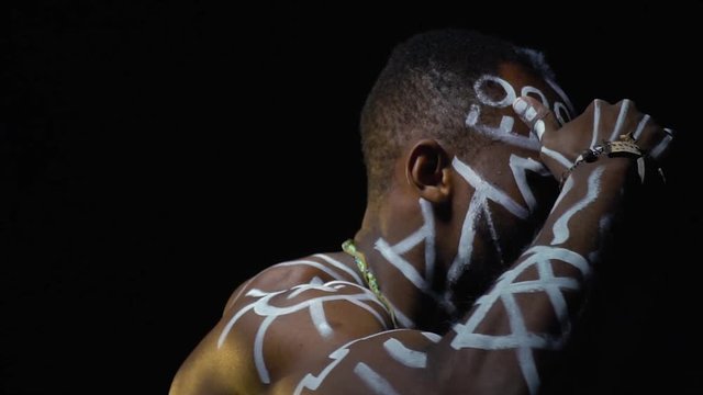 A black man in ritual patterns listens and looks out for someone in the dark