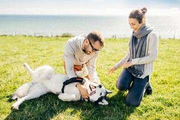 Fototapeta premium Young family couple have walk together with their pet, sits on green grass, have fun together. White husky enjoys love of hosts. Man and woman walk outside with dog. Leisure and people concept