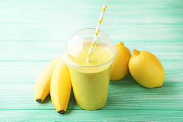 Yellow smoothie in plastic cup with bananas and lemons on mint wooden table