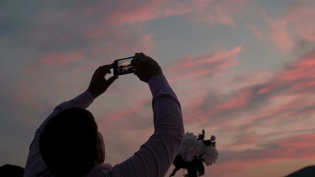 Man at dusk takes pictures of colorful sky on phone. Person stands with arms outstretched and makes shutter on device for memory of beautiful sunset.