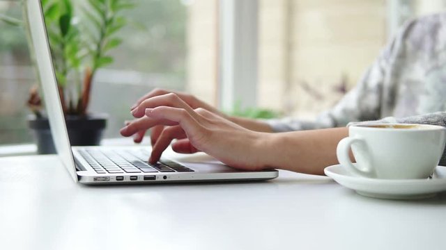 Closeup view of woman's hands working on laptop computer. Woman is sitting near the window in cafe and typing. White cup with coffee on the table