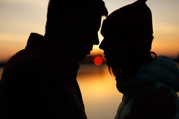 Silhouette of lovers couple on sunset by the lake. Sun between people profiles. Dusk sky, water on background. Passion in love concept, decoration, design for St. Valentines day.