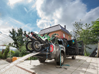 Young man securing two sports dirt bikes to the bed of the pickup truck using blue safety belt. © Georgii
