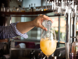 Bartender's hand holding a botlle with alcohol drink and puring it into the orange cocktail.
