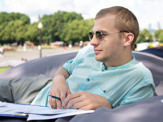Thoughtful young man in sunglasses laying on a cushion with paperwork outdoors and thinking.