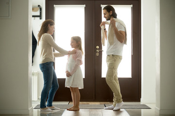Caring mother helping dressing little daughter for walk with father, happy family standing at home entrance door, loving mom and dad spending time taking care of kid girl, parents and child together