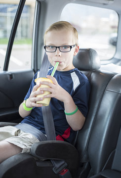 Cute boy sitting in a booster seat on a long car ride drinking a smoothie while safely strapped in a car seat. He is wearing his seatbelt and enjoying a drink.