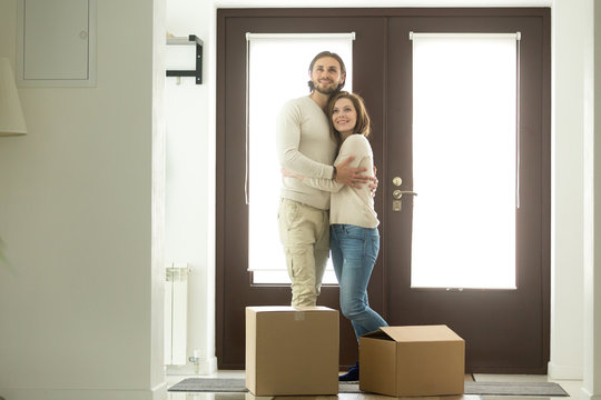 Moving day concept, excited young couple homeowners embracing in new home standing in modern hallway with cardboard boxes on floor looking around, mortgage loan, buying real estate and relocation