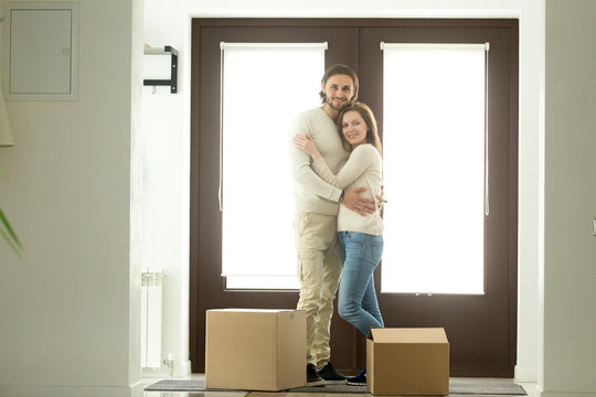 Happy young couple hugging looking at camera moving into new home, smiling homeowners embracing in hallway standing against door near cardboard boxes, affordable mortgage loan, buying house, portrait