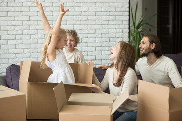 Happy child girl jumping out of cardboard box, family playing with kids in living room on sofa, parents and children laughing having fun together at moving day, move in new home or relocating concept
