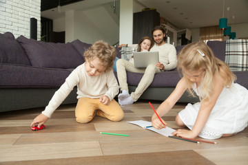 Kids siblings playing on floor while parents using laptop at home, preschool boy holding toy car, little girl drawing with colored pencils in album, family spending time together in living room