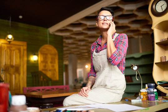Portrait of modern female artist wearing apron taking break from work sitting on table and speaking by phone in workshop - Powered by Adobe