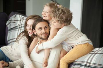 Happy parents and little children laughing having fun sitting on sofa, kids and wife hugging husband dad on couch, cozy loving family of four at home together, congratulating daddy with fathers day