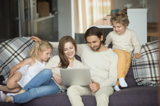 Happy family with kids using laptop sitting on sofa at home, smiling parents and son daughter relaxing on couch holding computer, man woman with children shopping together online in cozy living room