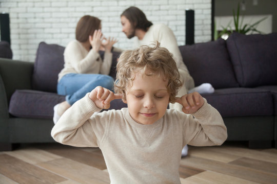 Little boy puts fingers in ears not to hear parents fighting at background, upset tired son suffering from mom and dad arguing, parental conflicts hurt kid, family divorce effect on children concept