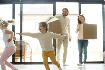 Kids playing while parents holding boxes coming into house on moving day, happy family with two children having fun move in big modern new home, couple son and daughter relocating with belongings