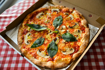Above view of tasty italian piza in openined paper box, on table with white and red checked blanket. Cozy restaurant. Pizza with basil, tomato cheese, champinions and herbs.