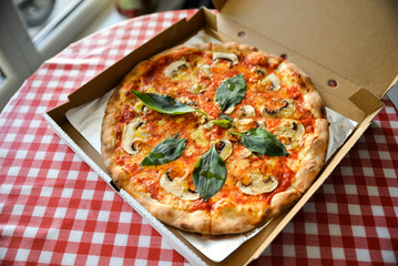 Above view of tasty italian piza in openined paper box, on table with white and red checked blanket. Cozy restaurant. Pizza with basil, tomato cheese, champinions and herbs.
