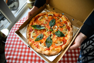 Crop of man holding paper box with italian pizza with cheese, basil, tomato, champinions. Food with option take away. Italian restaurant. 