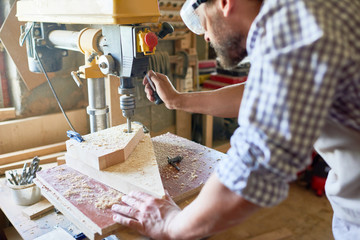 Mid section portrait of mature bearded carpenter wearing protective goggles using drilling machine for piece of wood  in modern workshop