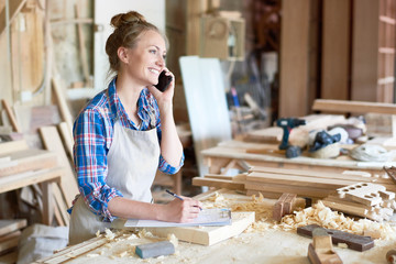 Portrait of modern young woman speaking by phone with client smiling and making notes in woodworking shop
