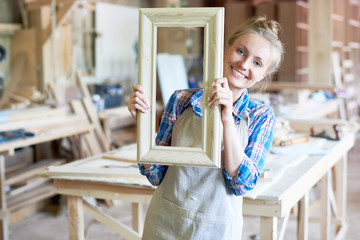 Portrait of happy young woman wearing apron smiling to camera holding wooden frame in modern...