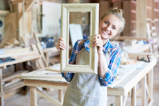 Portrait of happy young woman wearing apron smiling to camera holding wooden frame in modern joinery shop